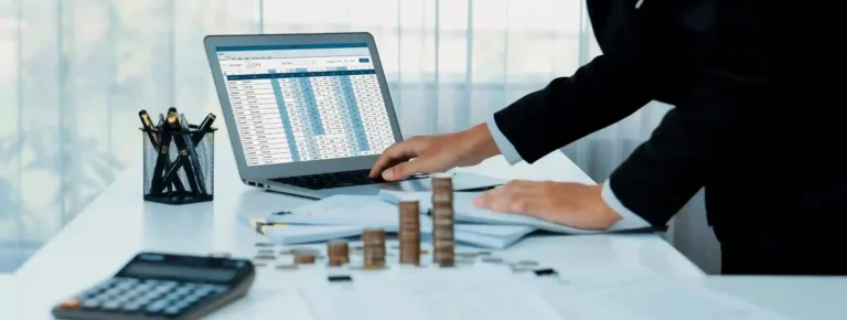 A professional accountant in a suit managing financial spreadsheets on a laptop next to stacked coins and a calculator. As an expert accounting firm in Dubai, Kloudac ensures your business remains compliant with all accounting services amidst a shifting regulatory landscape.